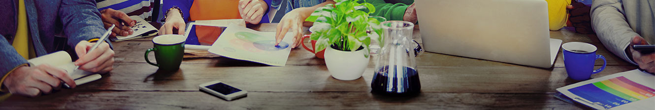 A group of people sitting at a table. There is a note pad, coffee cup, graphs and a laptop on the table.